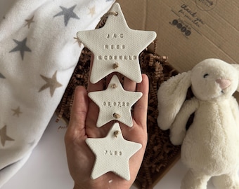 May include: Three off-white star-shaped ornaments with embossed text and twine hangers. The top star reads "JAG REES MCDONALD", the middle star displays "09.07.22", and the bottom star shows "7083". A plush white bunny and a star-patterned blanket are in the background.