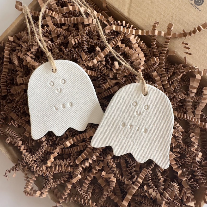 May include: Two white, ghost-shaped ornaments made of a textured material, each featuring a simple impressed face and the names LEO and OTIS. Each ornament hangs from a thin, natural-colored twine loop, designed for decorative hanging purposes.