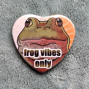 May include: A heart-shaped button with a cartoon frog face and the text "frog vibes only" in black and white.