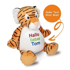 Peut inclure: Une peluche tigre avec des rayures orange et noires, des oreilles blanches et un ventre blanc. Le ventre présente un texte brodé en rouge, vert et bleu indiquant "Hallo lieber Tom". Un graphique circulaire orange avec le texte "Jeder Text Ihrer Wahl" est présent.