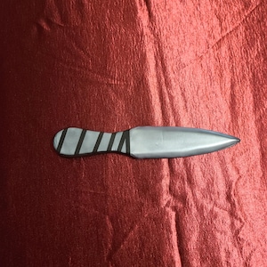 May include: A silver throwing knife with a pointed blade and a handle wrapped with black bands. The knife rests on a textured, red surface.