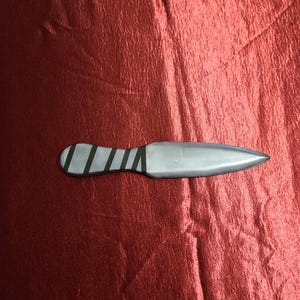 May include: A silver throwing knife with a pointed blade and a handle wrapped with black bands. The knife rests on a textured, red surface.