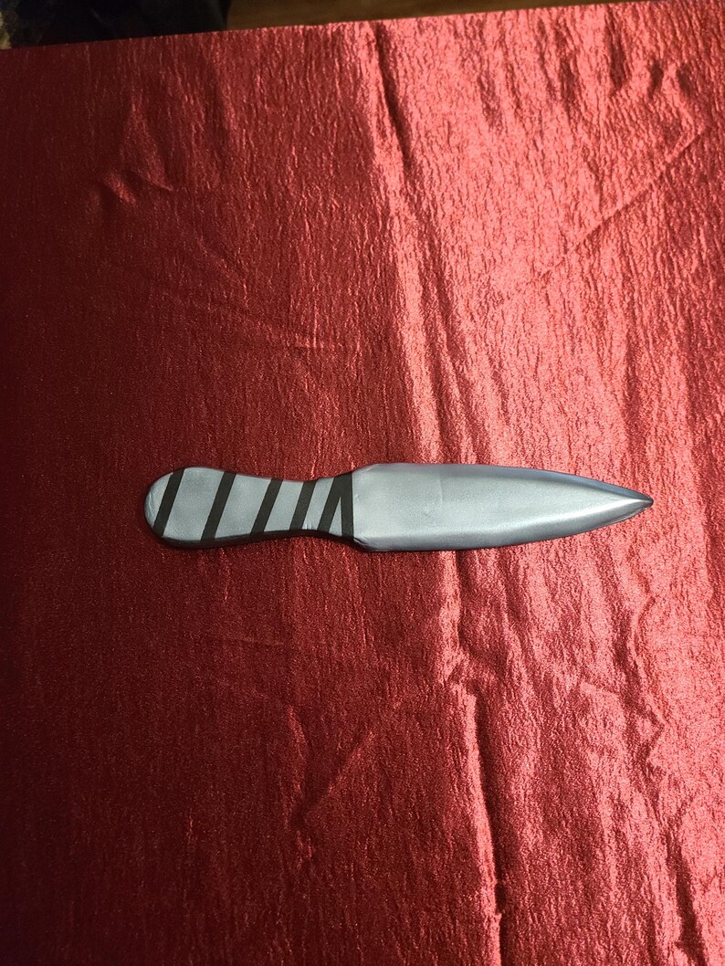 May include: A silver throwing knife with a pointed blade and a handle wrapped with black bands. The knife rests on a textured, red surface.