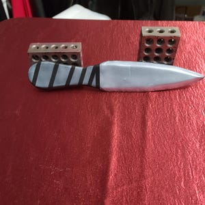 May include: A silver knife with a wrapped handle and two metal blocks with drilled holes. The knife has a pointed blade and is lying on a red surface. The handle is wrapped with black tape.