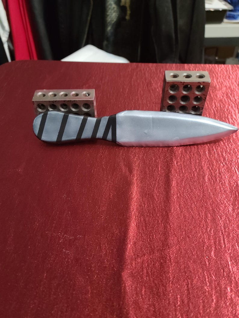 May include: A silver knife with a wrapped handle and two metal blocks with drilled holes. The knife has a pointed blade and is lying on a red surface. The handle is wrapped with black tape.