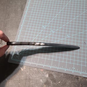 May include: A black letter opener, around 23 cm long, with a pointed tip, rests on a grey cutting mat. The handle is dark, and the blade is a lighter shade of black. The cutting mat features grid lines and centimetre measurements.