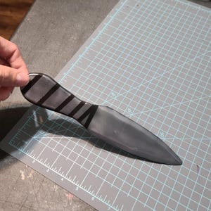May include: A black and grey dagger with black stripes on the handle. The dagger is on a grey cutting mat with blue grid lines and measurement markings.
