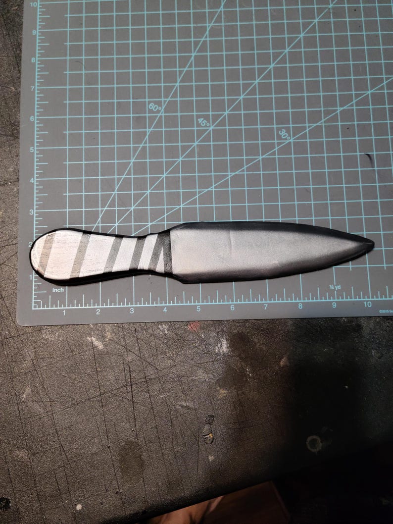 May include: A silver-coloured throwing knife with a striped handle, approximately 23 cm long. The blade is a solid, matte silver, and the handle has alternating silver and black stripes. The knife is lying on a grey cutting mat.