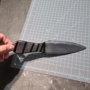 May include: A black throwing knife with a pointed tip and a handle decorated with diagonal black stripes. The knife is lying on a grey cutting mat with grid lines and centimetre measurements.