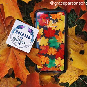 Op de afbeelding: Een sticker met de tekst "CREATED BY Kathleen CO." en een ontwerp van twee kwasten op een achtergrond van herfstbladeren. De sticker zit op een smartphone met een herfstbladerenpatroon als achtergrond. De tijd op de telefoon is 4:35.