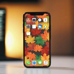 Op de afbeelding: Een zwarte iPhone met een herfstthema-achtergrond met kleurrijke esdoornbladeren. Het scherm van de telefoon toont verschillende app-pictogrammen, waaronder Camera, Klok, Agenda en Snapchat.