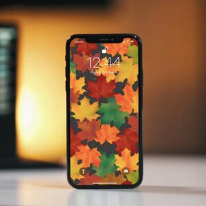 Op de afbeelding: Een zwarte iPhone met een herfstthema-achtergrond met kleurrijke esdoornbladeren. De tijd is 12:44 en de datum is vrijdag 4 oktober.