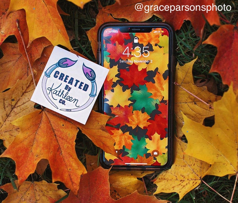 Op de afbeelding: Een sticker met de tekst "CREATED BY Kathleen CO." en een ontwerp van twee kwasten op een achtergrond van herfstbladeren. De sticker zit op een smartphone met een herfstbladerenpatroon als achtergrond. De tijd op de telefoon is 4:35.
