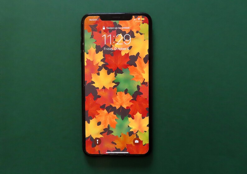 Op de afbeelding: Een zwarte smartphone met een vergrendelscherm waarop de tijd 11:29 en de datum vrijdag 6 oktober wordt weergegeven. De achtergrond is een patroon van kleurrijke herfstbladeren.