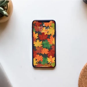 Op de afbeelding: Een smartphone met een blauwe hoes en een herfstthema wallpaper met kleurrijke bladeren. De tijd op het scherm is 12:44.