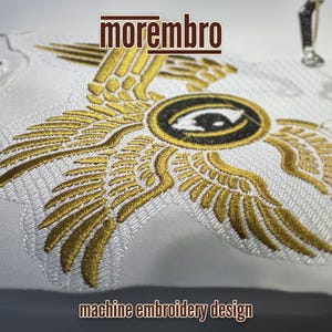 May include: Machine embroidery design featuring a pair of golden wings with an eye in the center. The design is on a white fabric and the text "morembro" is above the design. The text "machine embroidery design" is below the design.
