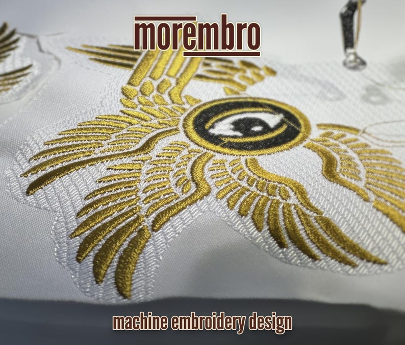 May include: Machine embroidery design featuring a pair of golden wings with an eye in the center. The design is on a white fabric and the text "morembro" is above the design. The text "machine embroidery design" is below the design.