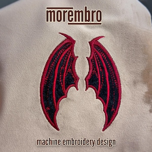 May include: Machine embroidery design featuring a pair of black wings with red outlines. The design is on a beige fabric and includes the text "morembro" and "machine embroidery design".