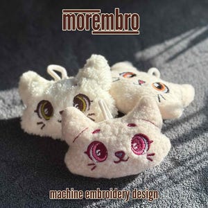 May include: Three white plush cat toys with embroidered faces. The cats have pink and white eyes and pink noses. The toys are on a grey textured surface.