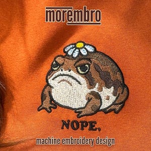 May include: An orange sweatshirt with an embroidered frog wearing a daisy crown. The frog has a grumpy expression and the word "NOPE" is stitched below it. The text "machine embroidery design" is also included.