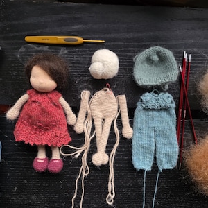May include: A doll in progress, with a red dress, blue pants, and a hat. Knitting supplies include yarn, needles, and a crochet hook. The doll's body is beige, with brown hair and pink shoes. A craft project.