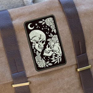 May include: A canvas messenger bag in a neutral tan color with dark brown leather straps and gold-colored metal buckles. A rectangular patch with a black background features two skulls, roses, stars, and a crescent moon.