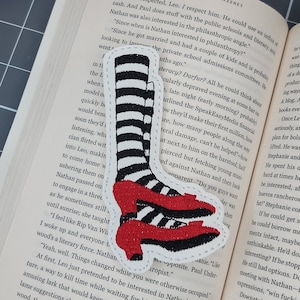 May include: Embroidered patch featuring the iconic ruby red slippers and striped socks from The Wizard of Oz. The patch has a white border and is placed on an open book. The shoes are red with a black heel.