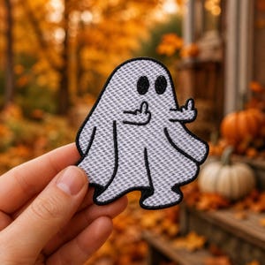 May include: A white embroidered patch shaped like a ghost with black outlines and black eyes. The ghost figure has two arms raised, each extending a middle finger. The patch features a textured, woven fabric appearance with a distinct black border.