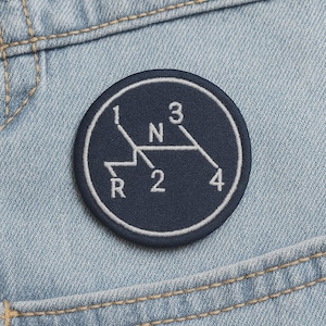 May include: A navy blue, circular patch with a white embroidered gear shift pattern. The patch features the numbers 1, 2, 3, and 4, along with "N" and "R" for reverse, all within a white border. The patch is set against a light blue denim background.