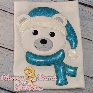 May include: White fabric with an embroidered polar bear wearing a blue glittery hat and scarf. The bear has black eyes and a gray nose. The text "Chevy Bomb Shoppe" is in the lower left corner.