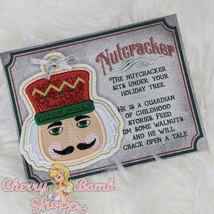 Può includere: Ornamento ricamato a forma di schiaccianoci con cappello rosso e oro glitterato, rifiniture verdi e baffi neri. L'ornamento è attaccato a un biglietto decorativo con la scritta "Nutcracker" e una poesia. L'ornamento è di "Cherry Bomb Shoppe".