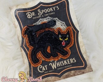May include: A black cat patch with orange glitter trim, set against a vintage-style label that reads "Dr. Spooky's Potions & Tinctures" and "Cat Whiskers." The cat has yellow eyes and an open mouth. The Cherry Bomb Shoppe logo is in the bottom left.