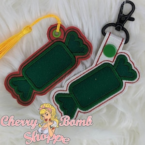 May include: Two candy-shaped keychains. One is red and green with a yellow tassel, the other is white and green with a black clip. Both have a green center and red trim, resembling wrapped candies. The logo "Cherry Bomb Shoppe" is in the bottom left.