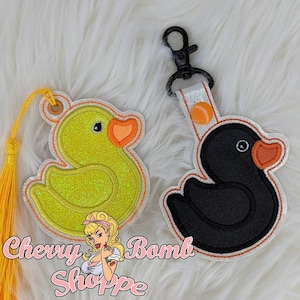 May include: Two duck-shaped keychains. One is yellow with orange accents and a yellow tassel. The other is black with orange accents and a silver strap with a black clip. The keychains are embroidered.