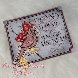 May include: Embroidered patch featuring a red cardinal bird with a red and white snowflake scarf, perched on a branch. The patch is on a gray background with the text "Cardinals Appear When Angels Are Near". The patch is from Cherry Bomb Shoppe.