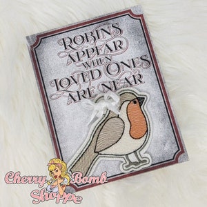 May include: A decorative sign with the text "Robins Appear When Loved Ones Are Near" in ornate lettering. A stitched robin with an orange breast is attached to the sign. The sign has a white ribbon and the Cherry Bomb Shoppe logo.