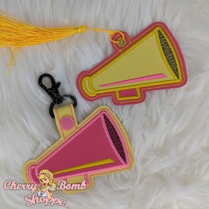 May include: Pink and yellow megaphone-shaped keychains with black accents and a yellow tassel. One keychain has a black clasp and a pink button. The keychains are embroidered with a yellow outline and the text "Cherry Bomb Shoppe".