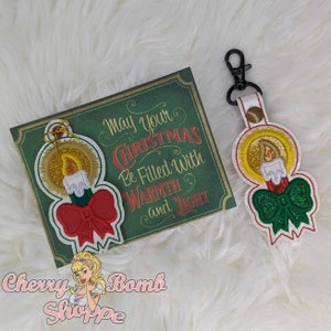 May include: A Christmas-themed set featuring a greeting card and a keychain. The card is green with gold trim and the text "May your Christmas Be Filled With Warmth and Light." The keychain and card both feature a candle design with a red bow and green accents.