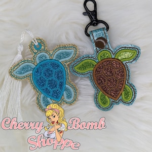 May include: Two glittery turtle-shaped accessories. One is a bookmark with a white tassel, the other a keychain with a black clasp. The turtles have blue, green, and brown accents with gold trim. The text "Cherry Bomb Shoppe" is at the bottom.