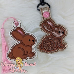 May include: Two embroidered bunny-shaped accessories. One is a bookmark with a pink tassel, the other is a keychain. Both have brown bodies, pink ears, and white outlines. The keychain has glitter accents and a black clasp.