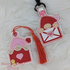 May include: Two Valentine's Day gnome-themed keychains. One keychain has a red envelope design with a pink heart and a gnome wearing a pink hat. The other keychain has a red tassel. The keychains are attached to a white strap with a black clasp.