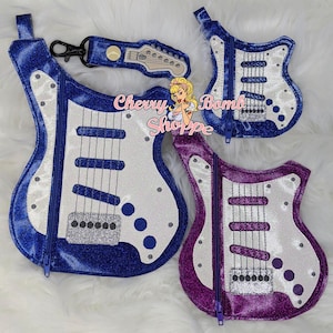 Könnte beinhalten: Eine Sammlung von gitarrenförmigen Reißverschlusstaschen und einem passenden Schlüsselanhänger. Die Taschen sind aus blauem und lila Glitzerstoff mit weißen und silbernen Akzenten gefertigt. Der Schlüsselanhänger hat ein Gitarrenkopf-Design.