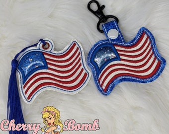 May include: Two patriotic American flag keychains. One has a blue tassel, the other a black clip. Both have red and white stripes, a blue glitter field of stars, and a white outline. The keychains are on a white fluffy surface.