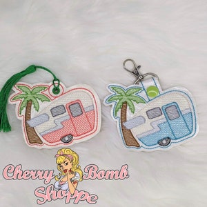 May include: Two embroidered camper keychains. One is red and the other is blue, each with a palm tree design. The keychains have a white border and a green tassel or a silver clip. The text "Cherry Bomb Shoppe" is at the bottom.