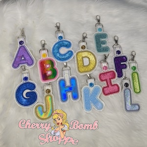 May include: A collection of glittery alphabet letter keychains in various colors, including purple, blue, gold, and pink. Each letter is outlined in white and attached to a silver clip. The "Cherry Bomb Shoppe" logo is visible at the bottom.