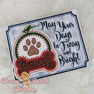 May include: A Christmas ornament featuring a dog paw print and a bone shape, with the text "May Your Days be Furry and Bright." The ornament has red, green, and brown accents, and is attached to a decorative card.
