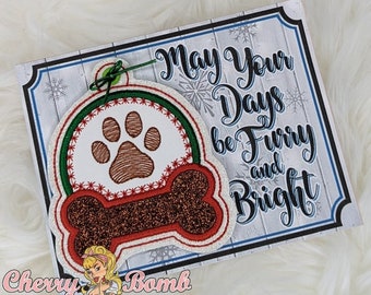 May include: A Christmas ornament featuring a dog paw print and a bone shape, with the text "May Your Days be Furry and Bright." The ornament has red, green, and brown accents, and is attached to a decorative card.