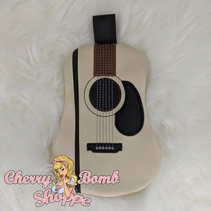 以下が含まれることがあります： クリーム色のギター型ポーチ。黒いジッパーと黒いストラップが付いています。ギターのデザインを模倣した茶色と黒のディテールが特徴で、弦やサウンドホールが含まれています。Cherry Bomb Shoppeのロゴが見えます。