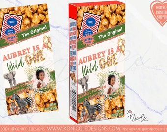 May include: A green, red, and brown cardboard box with a jungle animal theme. The box has a picture of a baby elephant, a zebra, a tiger, and a giraffe. The text on the box reads "Aubrey is a Wild One" and "Caramel Popcorn & Peanuts". The box is 1 oz (28.35 g).