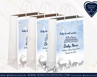 Könnte beinhalten: Drei weiße Papier-Geschenkbeutel mit blauen und silbernen Schneeflocken-Designs. Die Beutel haben braune Henkel und sind mit einem Rentier und anderen Wintertieren verziert. Der Text auf den Beuteln lautet "Baby it's cold outside... Thank you for welcoming Baby Nova Aubrey & Chris Baby Shower December 24, 2022".